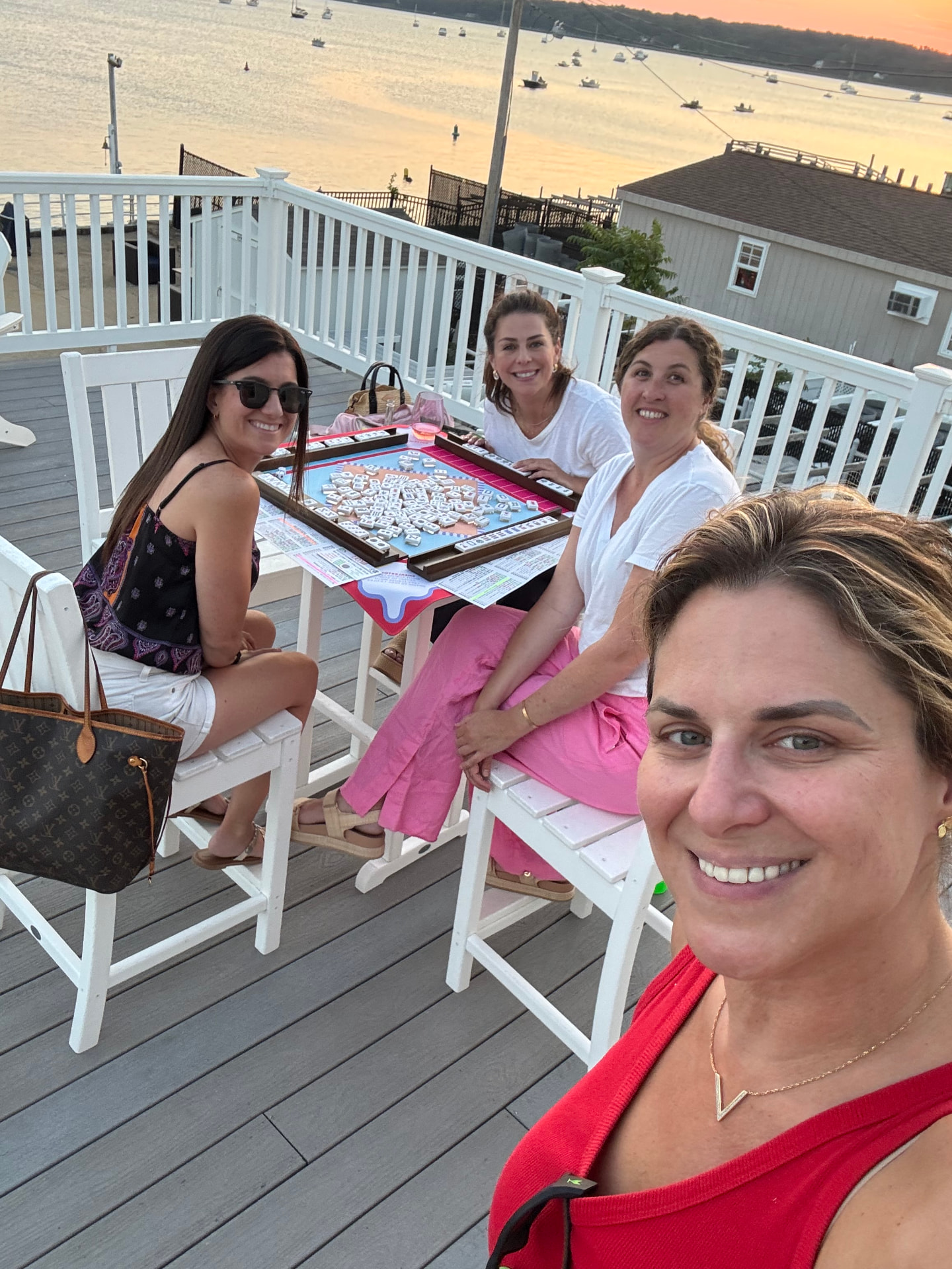 Four friends playing mahjong on a waterfront deck at sunset, Sandra in red taking a selfie