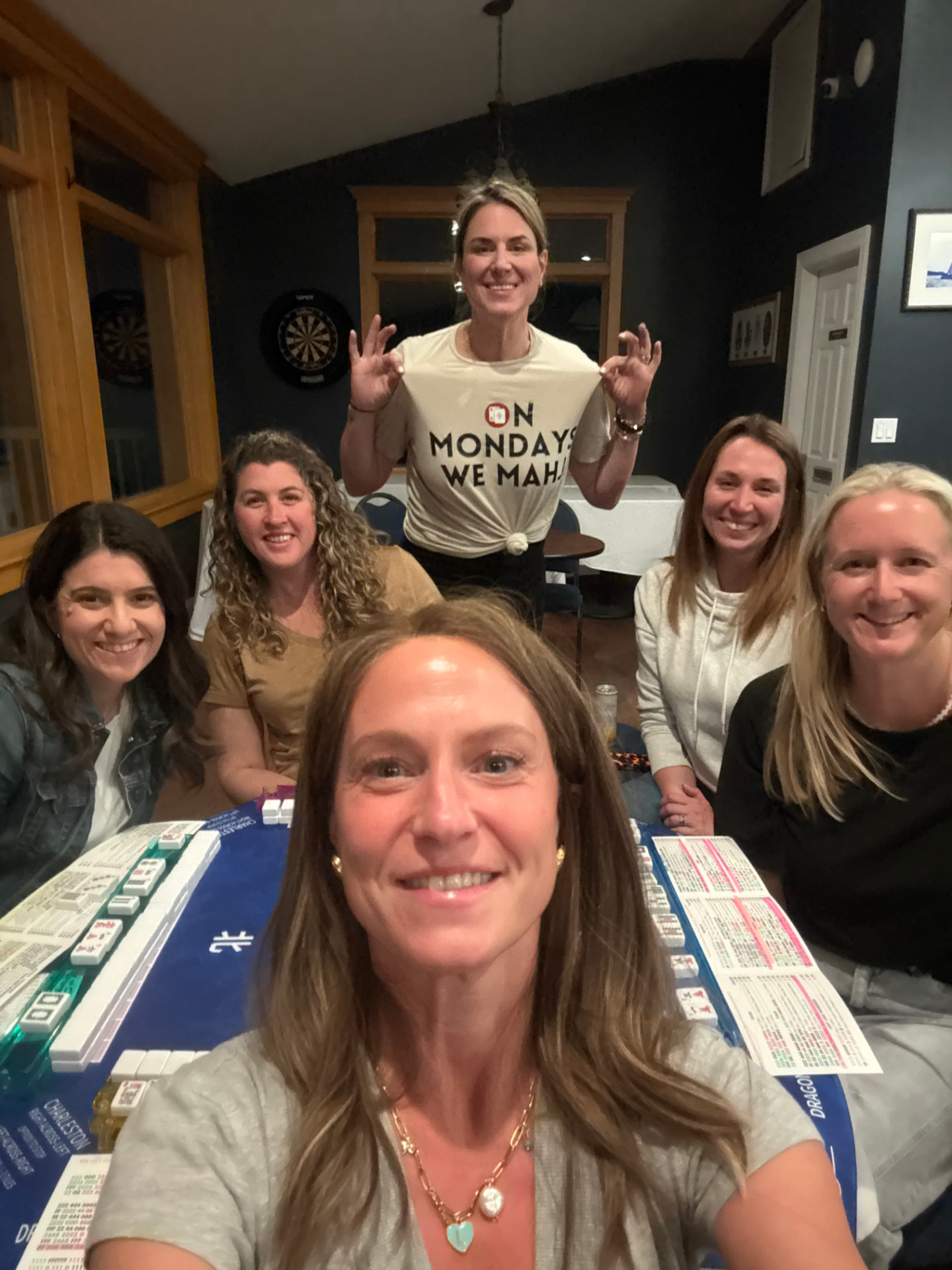 Six friends smiling around a mahjong table with a blue mat and loaded racks, one wearing an On Mondays We Mahj shirt