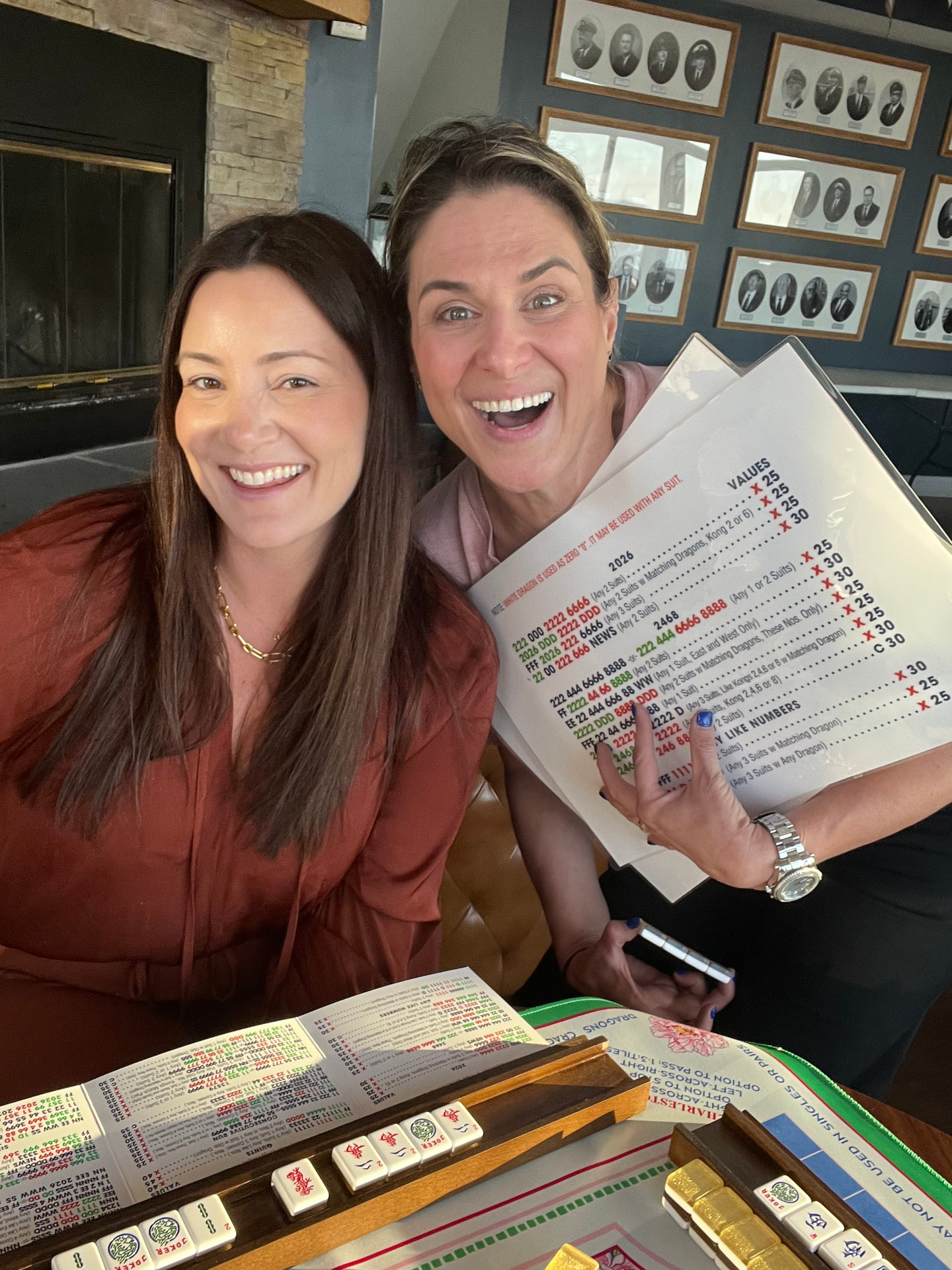 Two friends cheering as one holds up laminated mahjong score cards above a table of gold-backed tiles