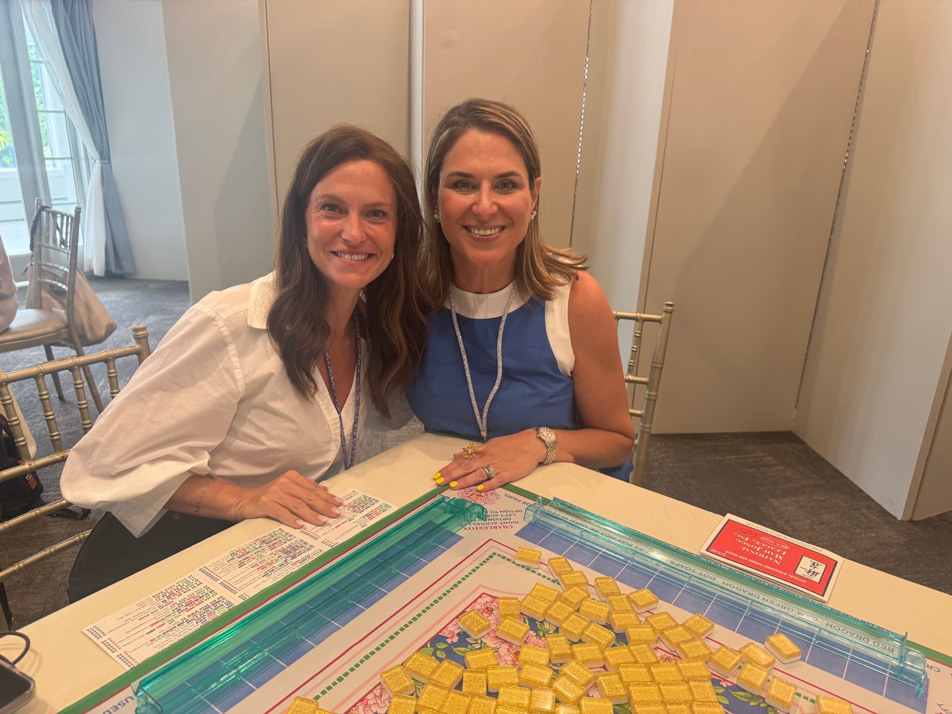 Sandra and a friend smiling at a mahjong table set with gold-backed tiles and score cards