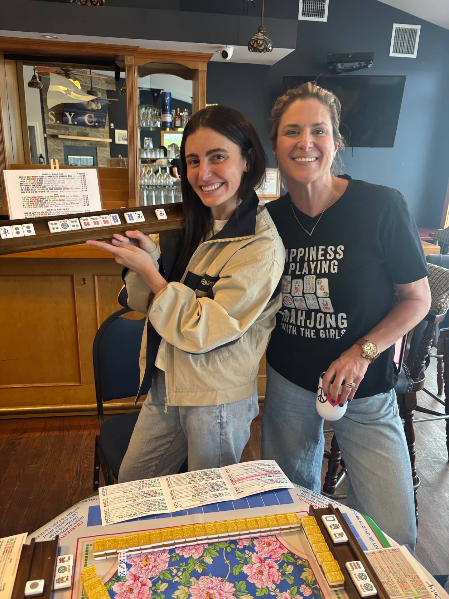 Two friends posing proudly at a mahjong table, one showing a full hand on a wooden rack, the other wearing a Happiness Is Playing Mahjong With The Girls shirt