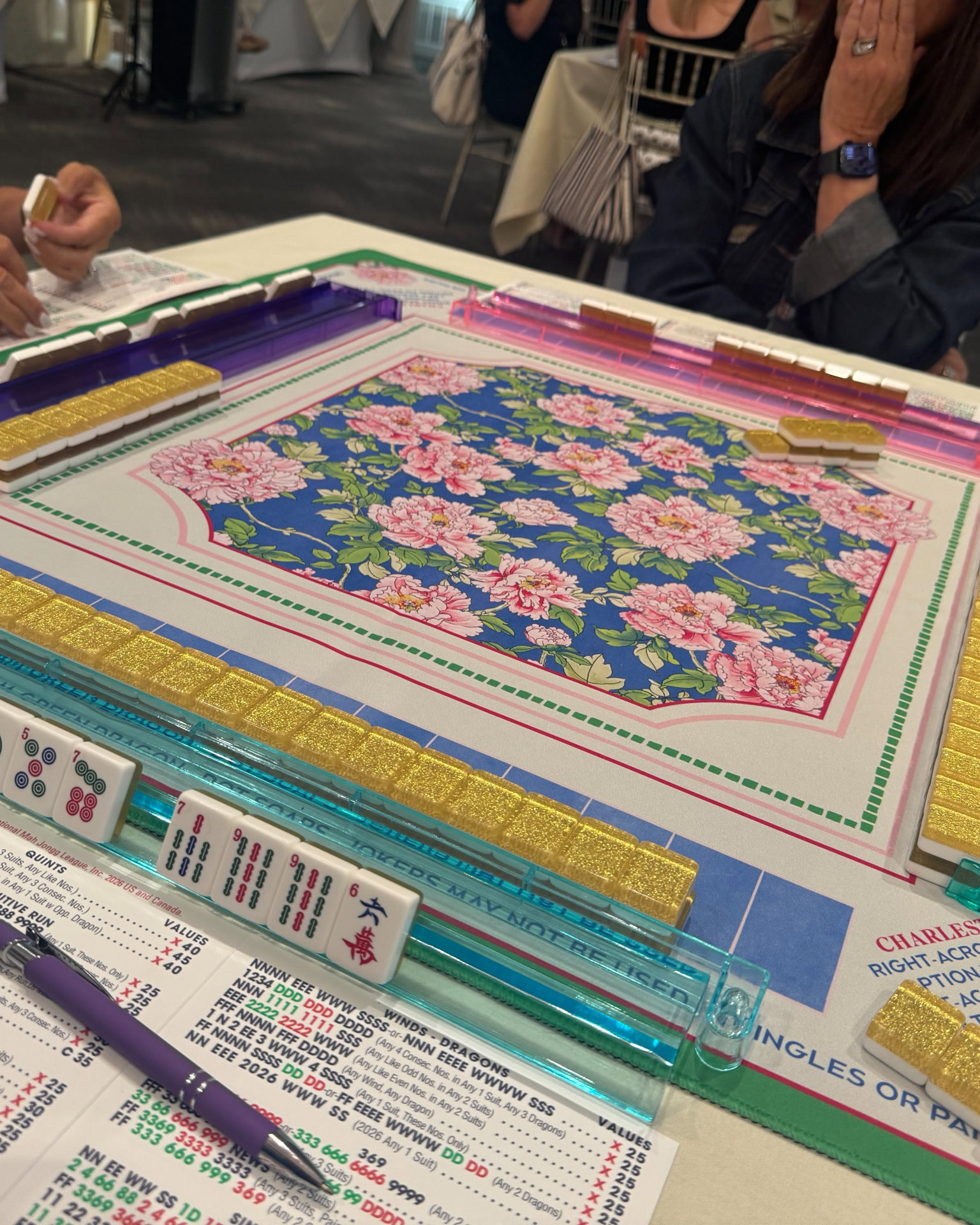 A real Mahjong table mid-game: gold glitter tiles on acrylic racks, the league card resting at the table's edge